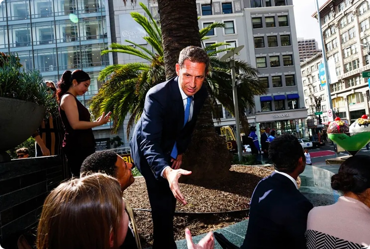 Man in a suit speaking with a group of people in a downtown San Francisco plaza