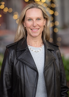 Professional headshot of a woman smiling outdoors, wearing a black leather jacket with blurred lights in the background