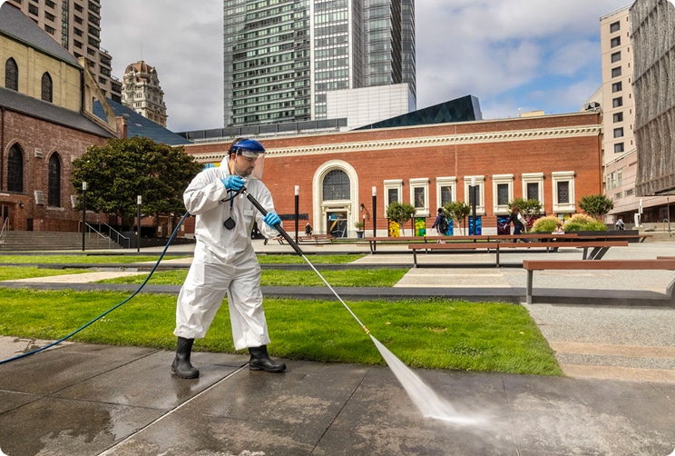 Worker in protective gear using a pressure washer to clean a sidewalk in a downtown San Francisco plaza